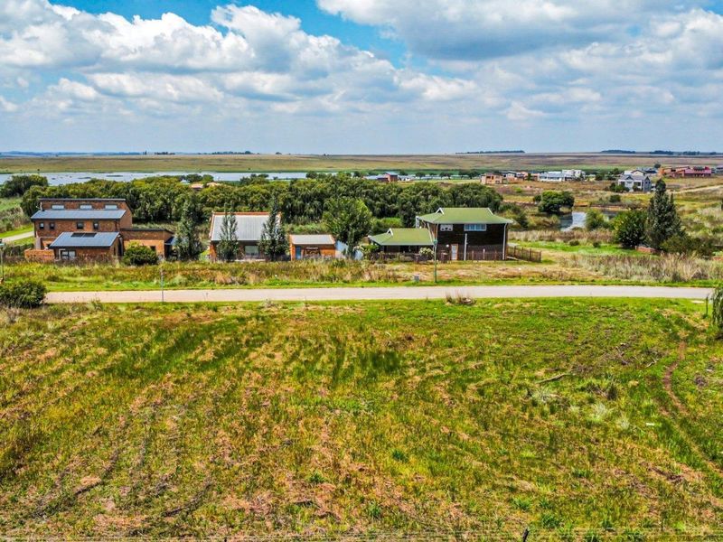 An aerial view of a vacant residential plot featuring open grassland with a paved road and surrounding neighborhood houses in the background.