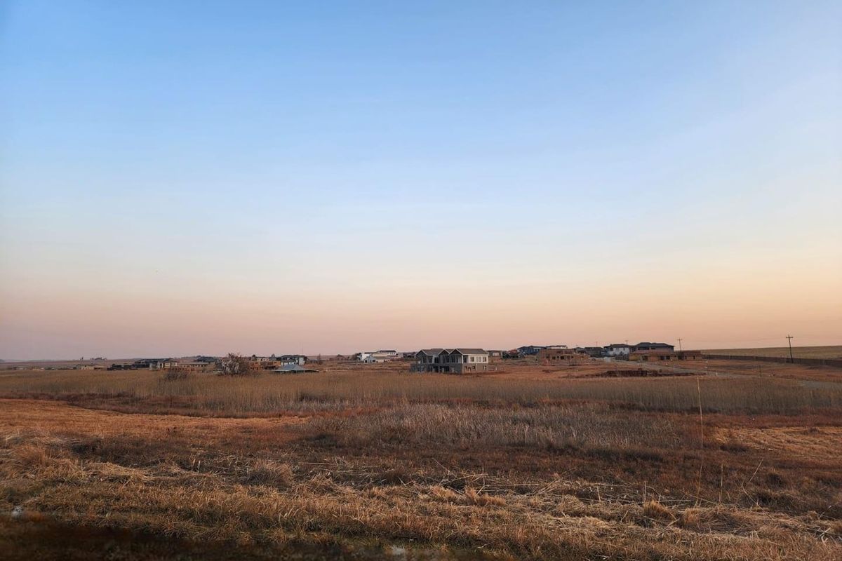 A wide-angle view of a vacant residential plot featuring dry grassland with distant suburban development visible on the horizon.
