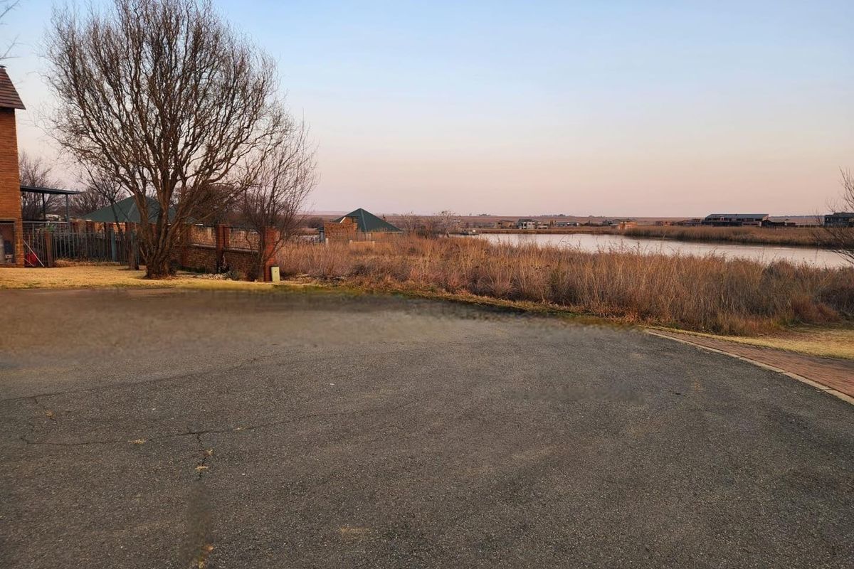 A view of a vacant residential plot featuring a paved driveway, natural vegetation, and scenic water frontage.