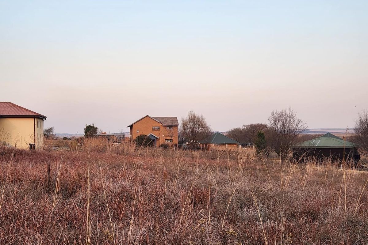 A view of a vacant residential plot featuring dry grass, with neighboring houses visible in the background.