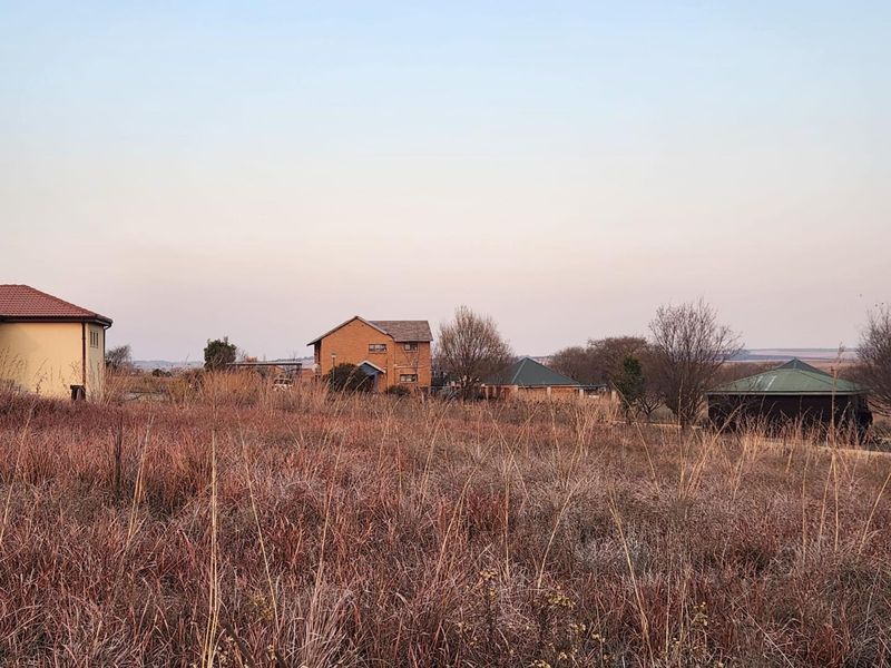 A view of a vacant residential plot featuring dry grass, with established neighboring houses visible in the background.