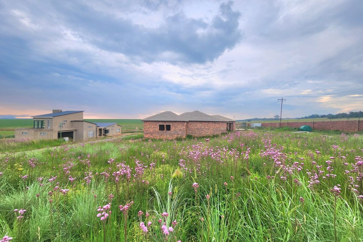 A view of a residential plot featuring an unfinished brick structure under construction, surrounded by open, undeveloped land.