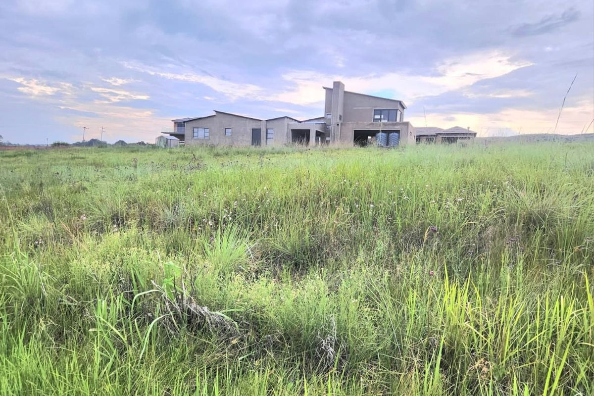 A view of an unfinished residential building structure set within a large, undeveloped grassy plot.