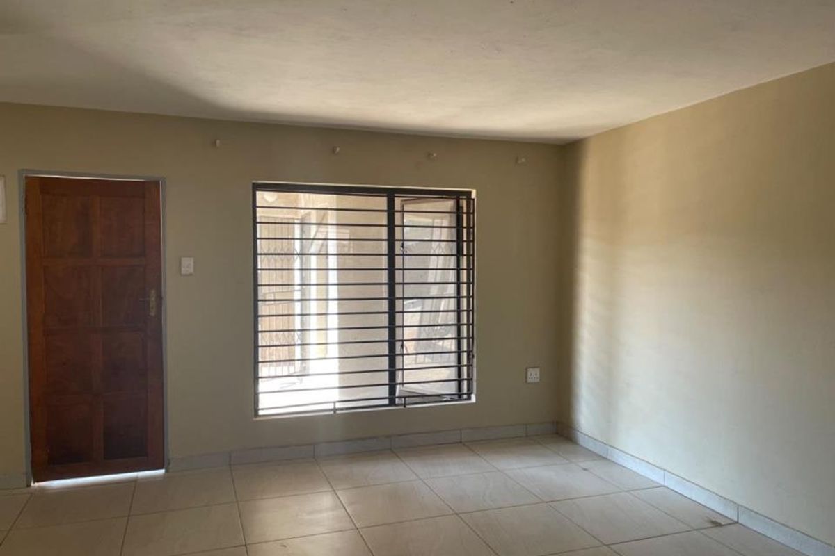 An empty living room featuring tiled flooring, a wooden entrance door, and a window fitted with security bars.