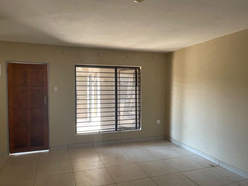 An empty living room featuring tiled flooring, a wooden entrance door, and a window fitted with security bars.