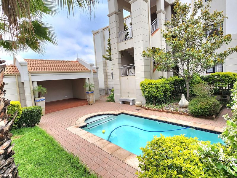A well-maintained communal pool area featuring a paved deck, covered patio space, and established garden landscaping within an apartment complex.