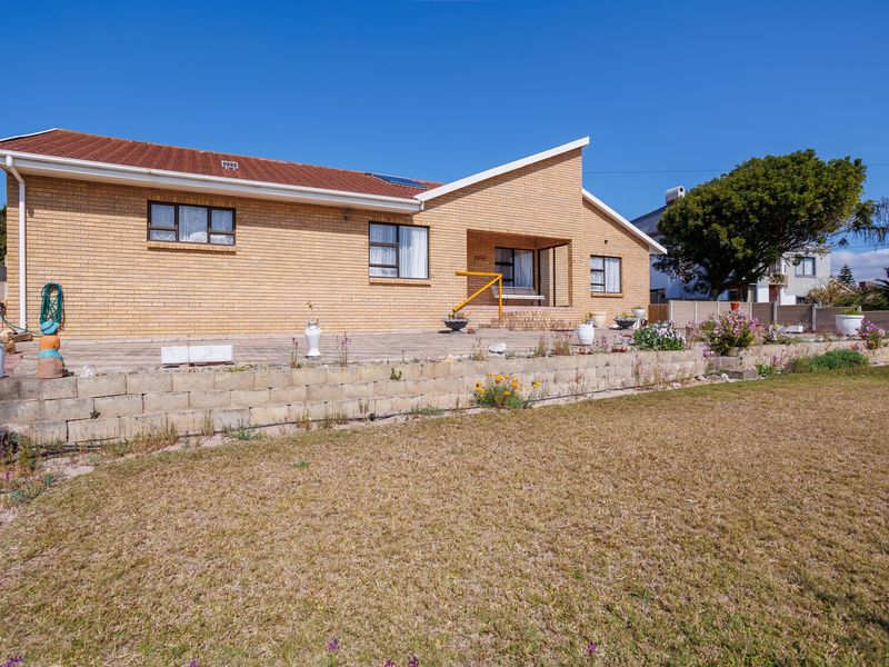 Rear view of a brick house featuring a paved patio area, a low retaining wall, and a spacious garden under a clear blue sky.