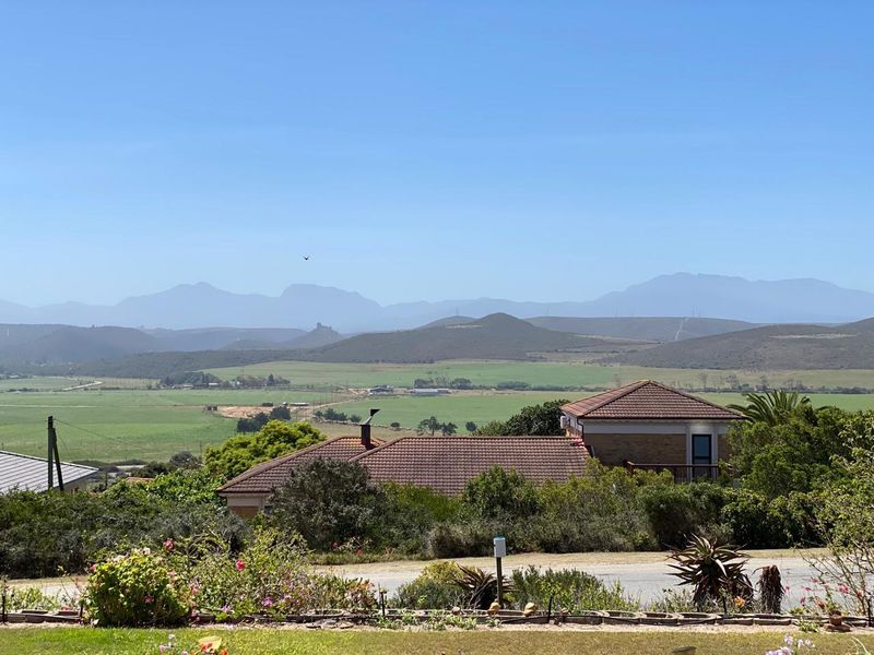 A scenic elevated view overlooking rolling green hills and distant mountains, featuring a residential property with a tiled roof in the foreground.