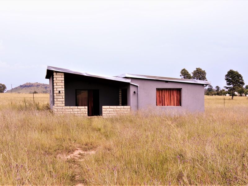 A detached house featuring a pitched roof, stone masonry accents, and a covered patio, set in a rural landscape with tall grass.