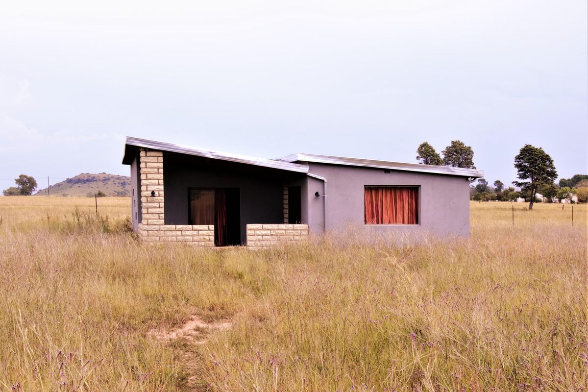 A detached house featuring a pitched roof, stone masonry accents, and a covered patio, set in a rural landscape with tall grass.