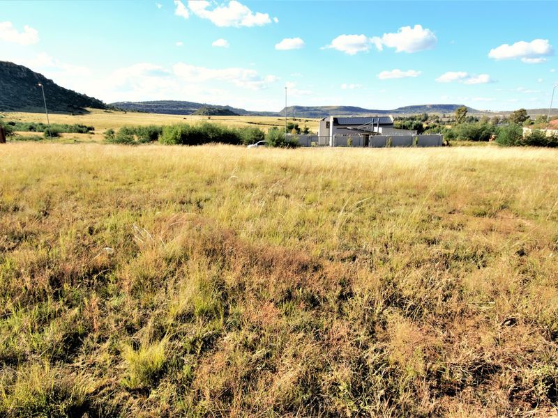 A wide-angle view of a vacant residential plot featuring natural grassland, with surrounding landscape and distant residential structures visible.