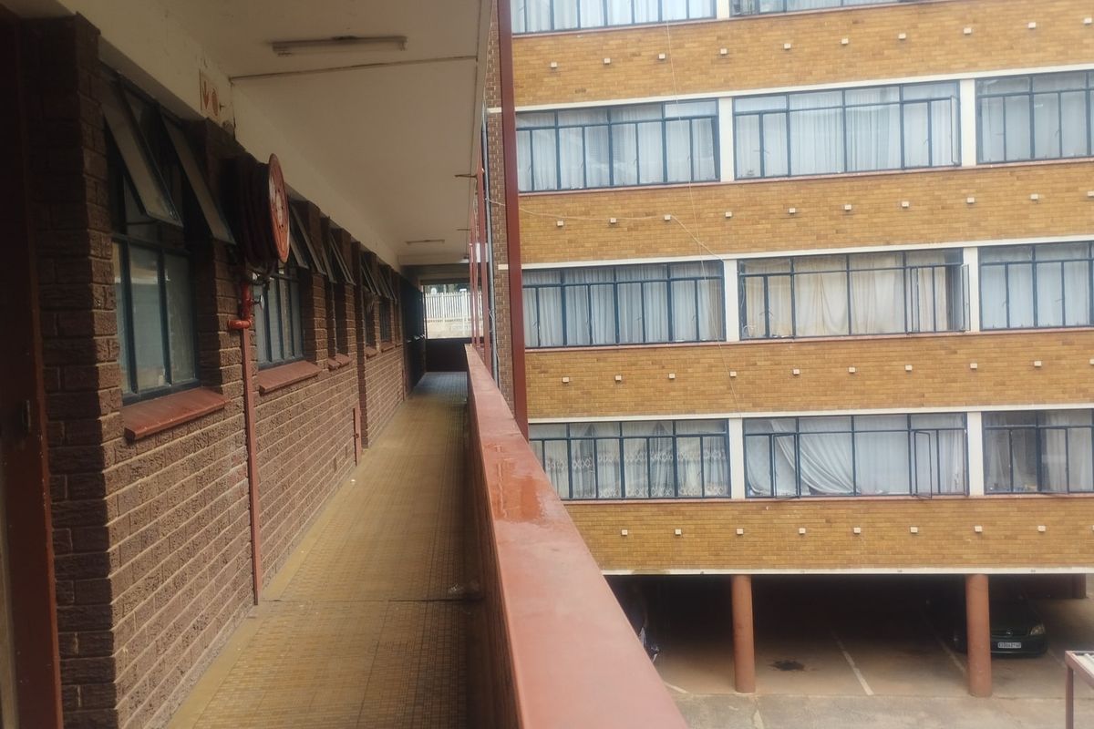 An exterior view of an apartment building featuring a covered walkway, brick facade, and steel-framed windows overlooking a parking area.