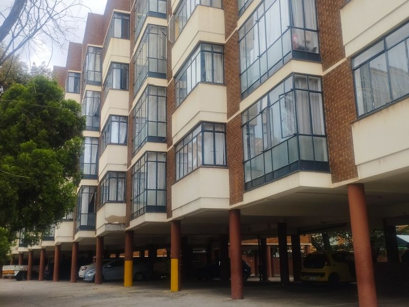 Exterior view of a multi-story apartment building featuring brick facade, enclosed balconies, and a ground-level covered parking area supported by concrete pillars.