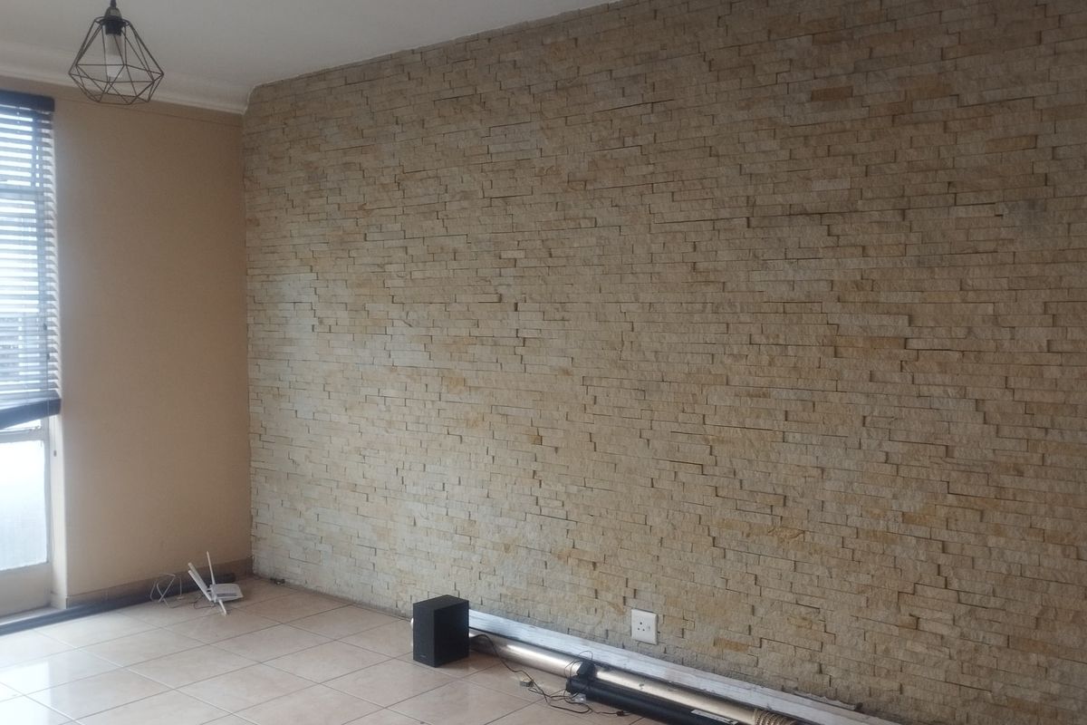 A living room featuring a prominent natural stone cladding feature wall, tiled flooring, and large windows with venetian blinds.