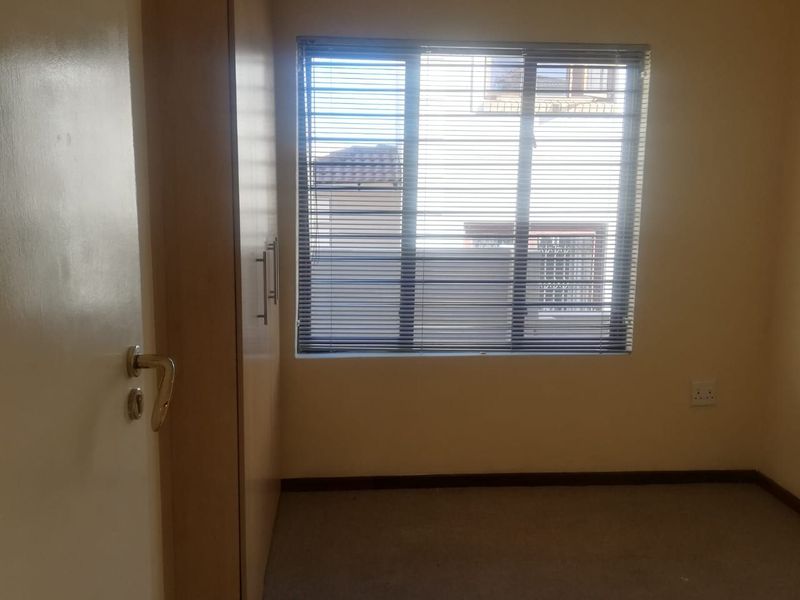 A simple, empty bedroom featuring built-in cupboards, carpeted flooring, and a window fitted with venetian blinds.