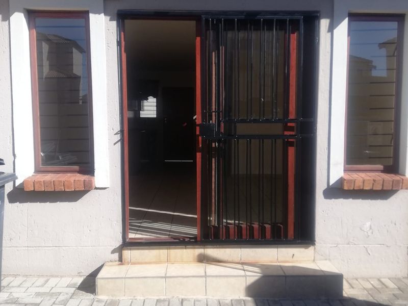 Front entrance of the townhouse featuring a security gate, tiled step, and brick-accented windows overlooking a paved driveway area.