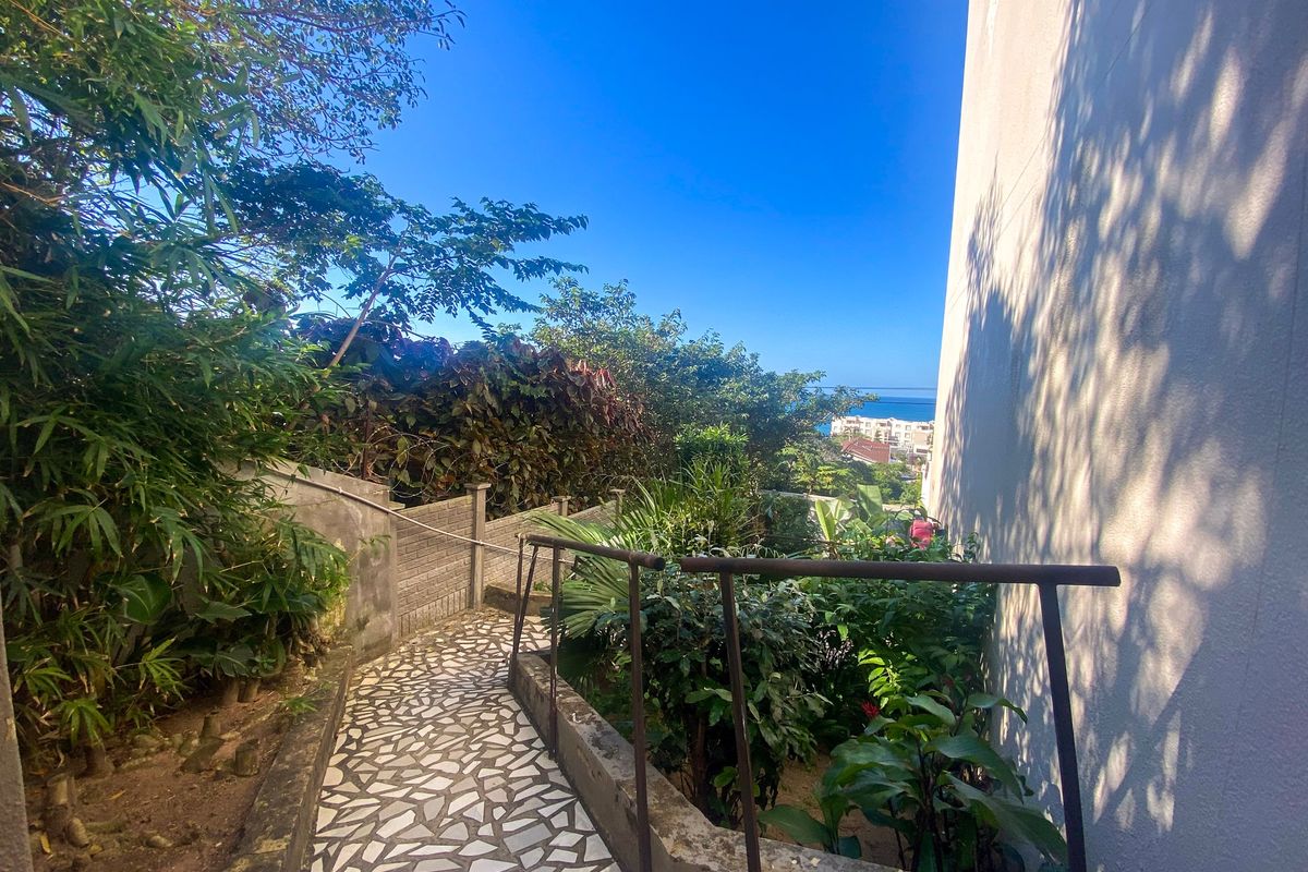 A paved outdoor pathway with metal handrails leading through a landscaped garden area, offering a glimpse of the ocean in the distance.