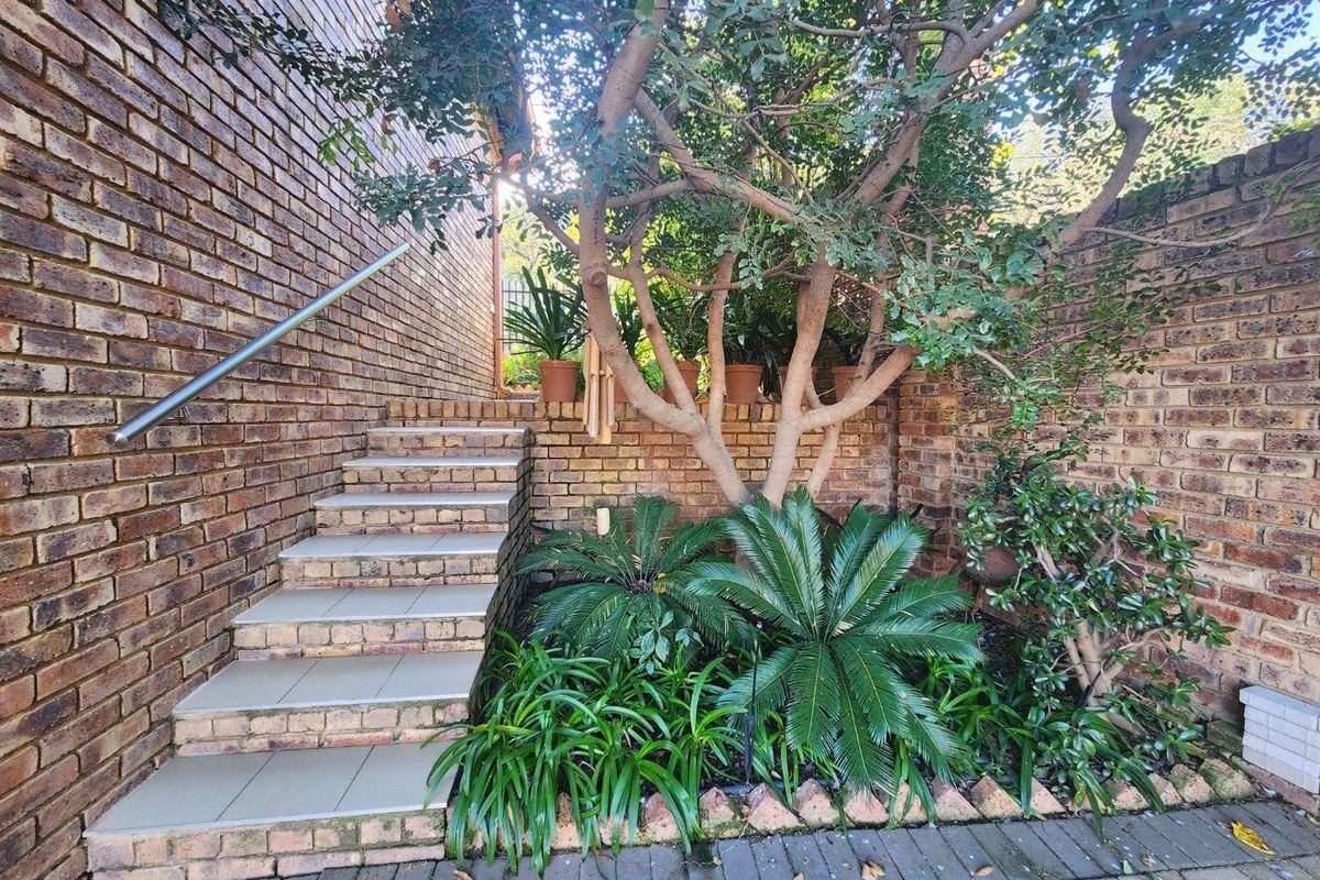 A well-maintained outdoor area featuring a brick staircase with a stainless steel handrail, leading to a landscaped garden bed with mature trees.