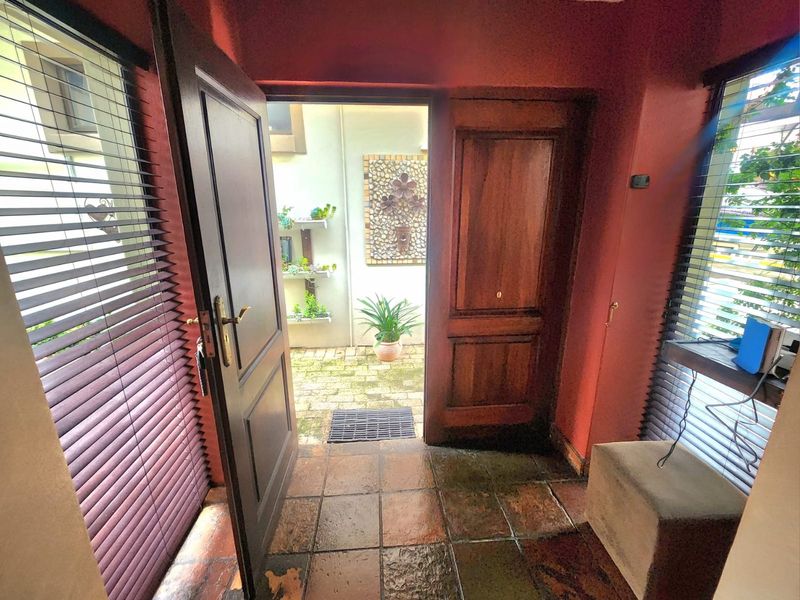 An entryway featuring terracotta-style floor tiles, dark wood doors, and venetian blinds, leading out to a small paved courtyard area.