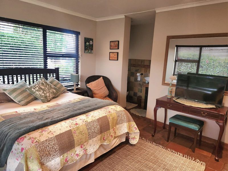 A bedroom featuring terracotta floor tiles, large windows with fitted blinds, and an open-plan ensuite bathroom with tiled walls.