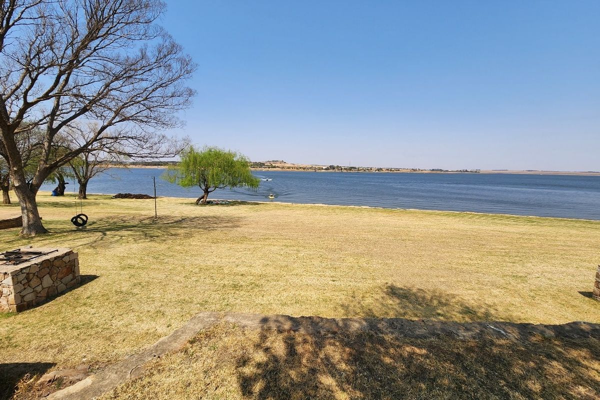 Expansive waterfront view overlooking a large dam with mature trees and a stone fire pit area on the grassy bank.