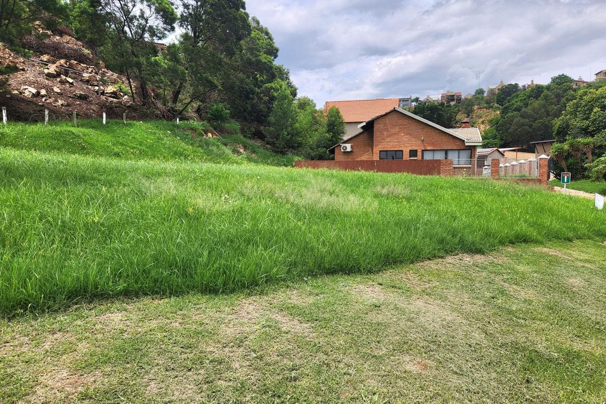 A vacant residential plot featuring a sloped grassy landscape with established trees and adjacent brick residential structures.