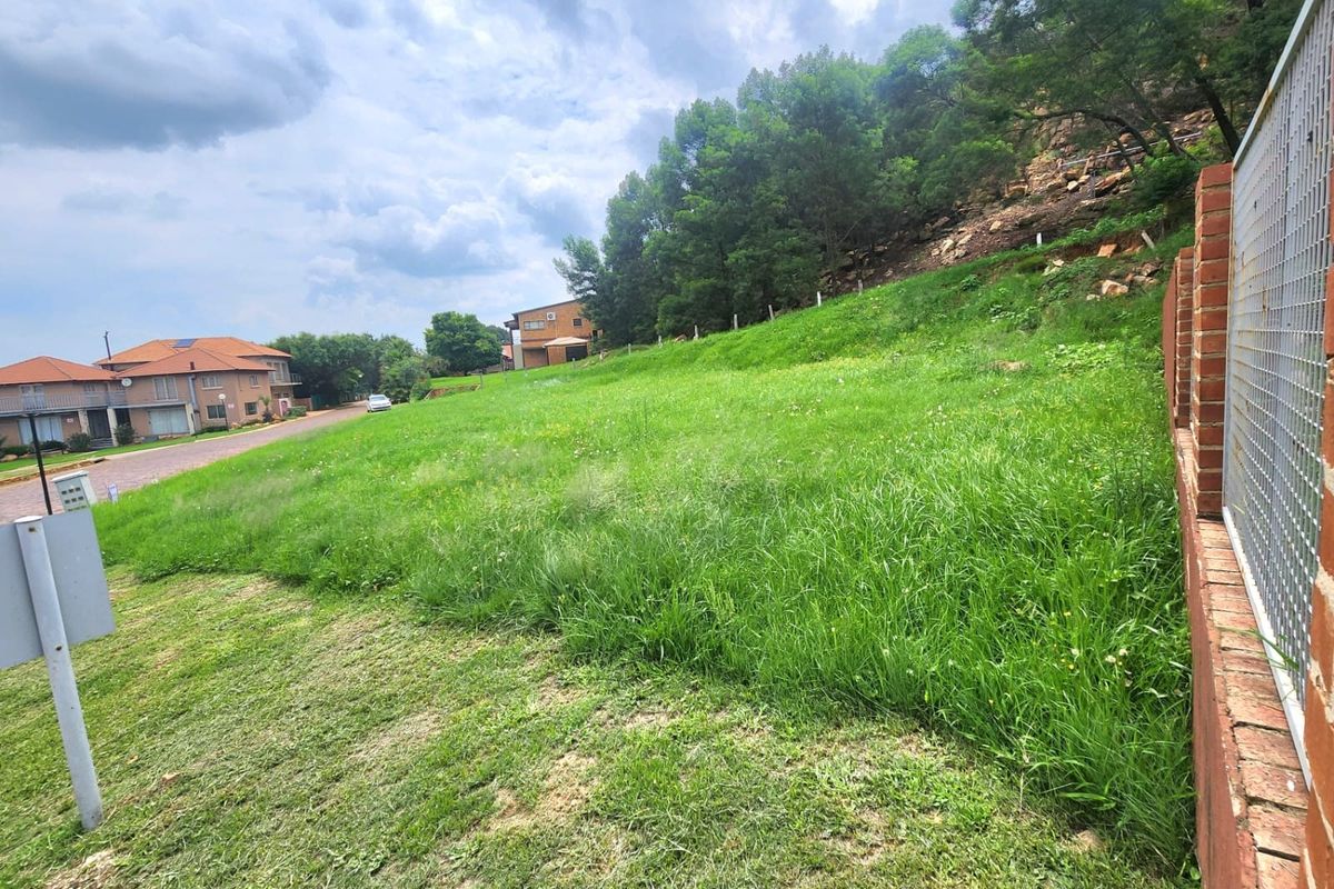 A vacant residential plot featuring a sloped grassy landscape, bordered by a brick and metal mesh boundary wall in a suburban setting.