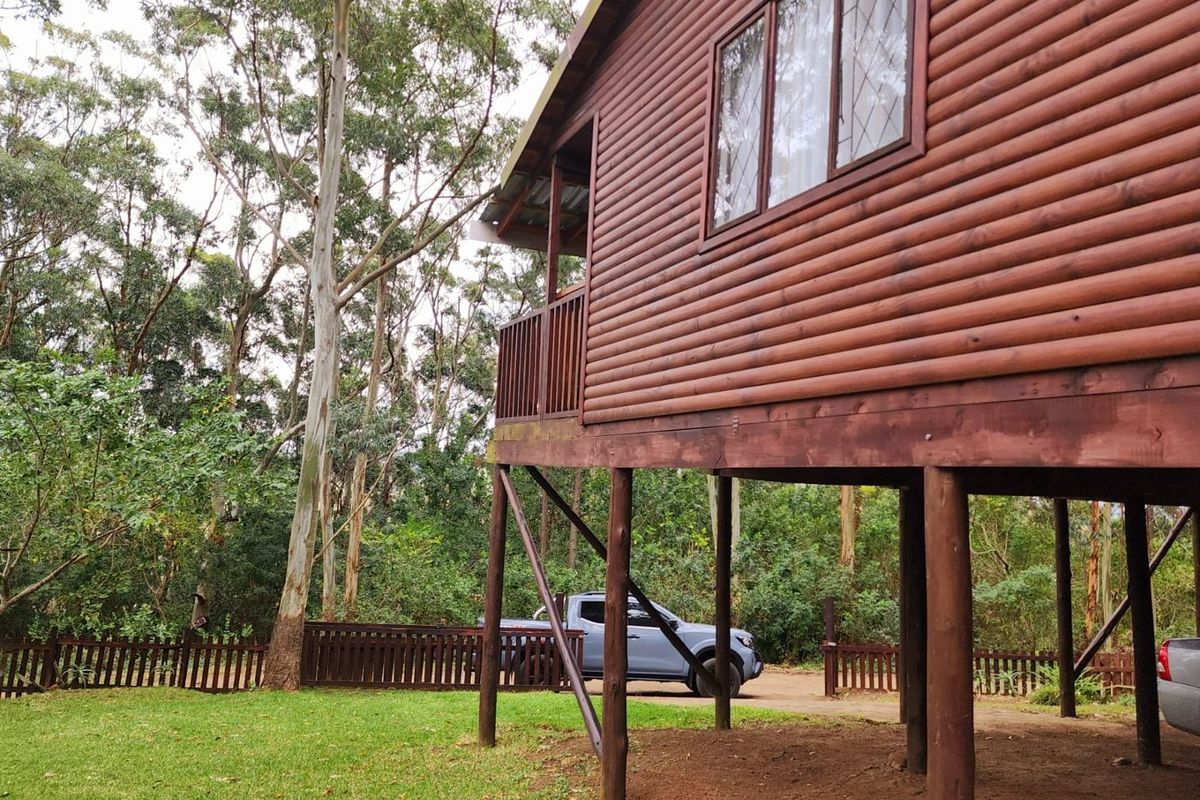 A raised timber log cabin townhouse featuring a balcony deck, supported by wooden stilts, set within a natural wooded garden environment.