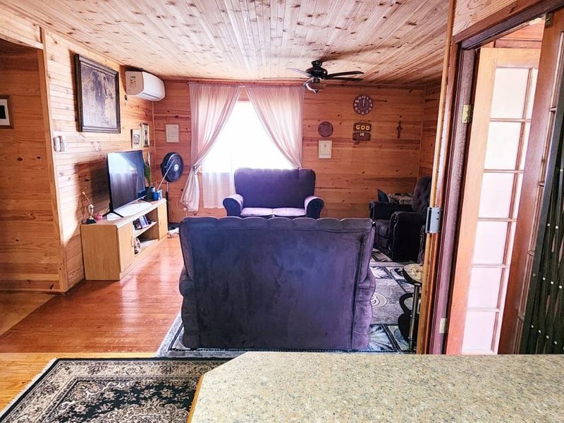 A cozy living room featuring extensive timber wall and ceiling cladding, timber flooring, a ceiling fan, and a wall-mounted air conditioning unit.