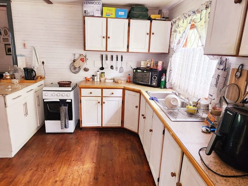 A dated kitchen featuring timber wall paneling, wooden flooring, white cabinetry, and a freestanding stove, offering potential for a modern renovation.