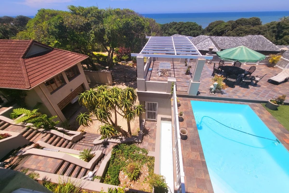 Aerial view of a coastal property featuring a swimming pool, tiled patio area with pergola, landscaped garden, and paved driveway.
