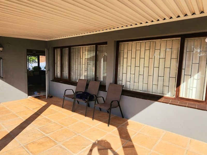 A covered patio area featuring tiled flooring, a corrugated metal roof, and security bars on the windows, providing a functional outdoor space.