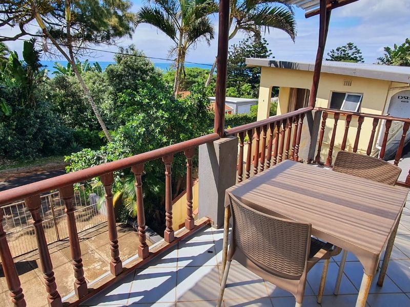 An elevated balcony featuring tiled flooring and a wooden balustrade, offering an outdoor space with views of the surrounding greenery and distant ocean.