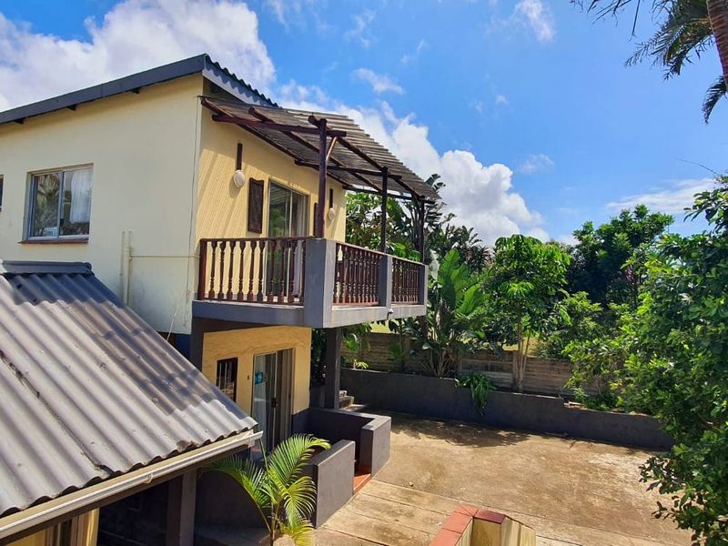 Rear view of the property featuring a two-story structure with an upper-level balcony, concrete patio area, and established garden surroundings.