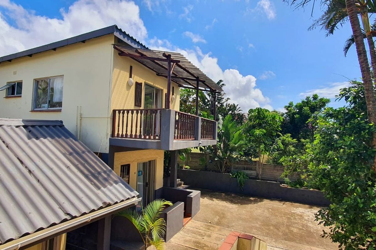Rear view of the property featuring a two-story structure with an upper-level balcony, concrete patio area, and established garden surroundings.