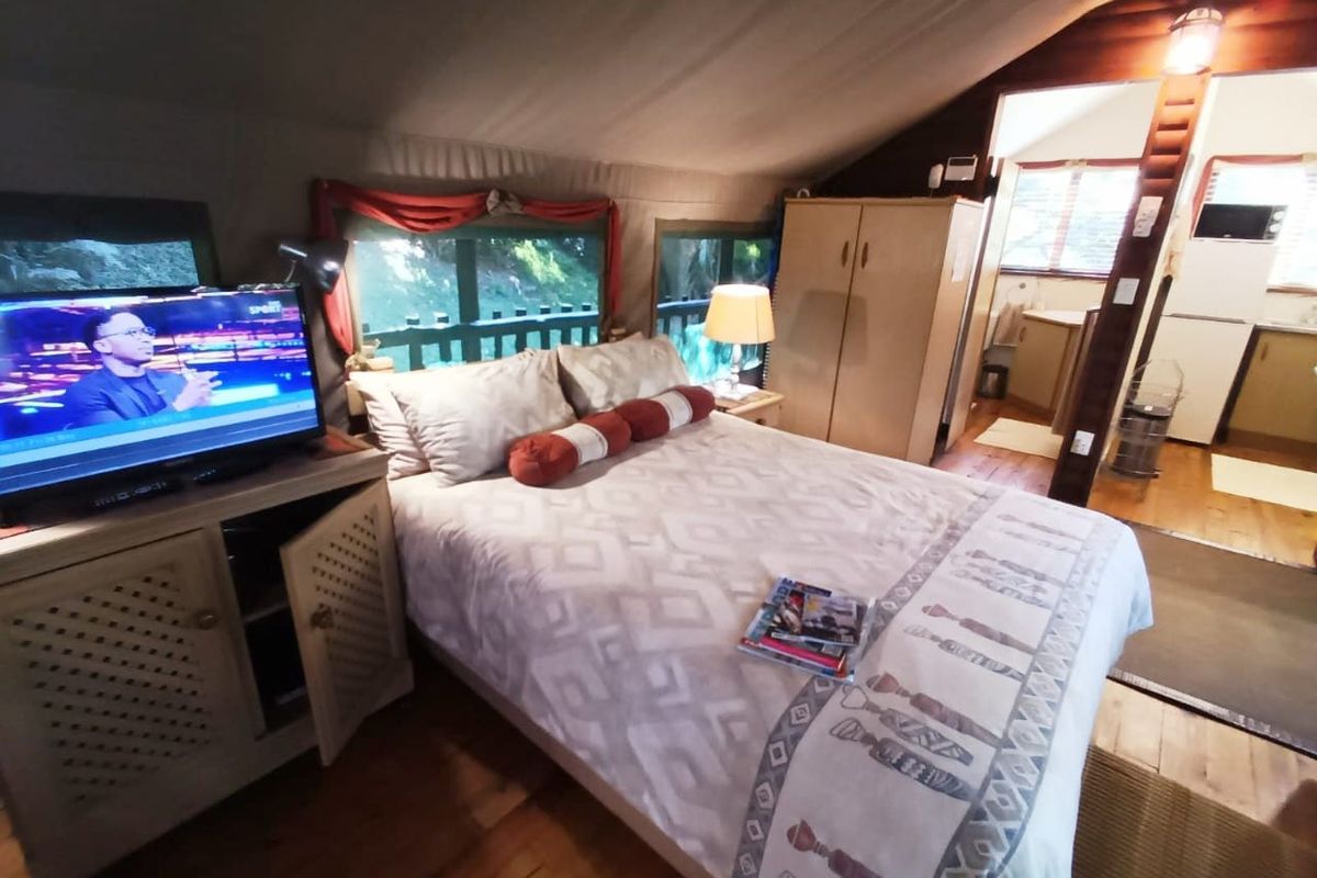 A rustic bedroom featuring timber flooring, canvas ceiling, and integrated wardrobe, with an adjoining kitchenette area visible in the background.