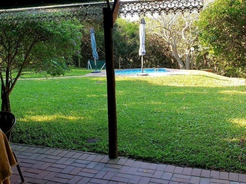 A covered patio area with brick paving and timber posts, overlooking a lush green garden and a swimming pool.