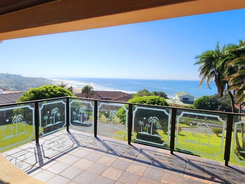A spacious covered balcony featuring tiled flooring and a decorative glass balustrade, offering expansive ocean and coastal views.