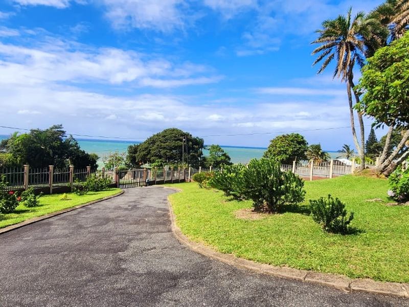 A well-maintained front driveway and landscaped garden area featuring mature trees, metal fencing, and scenic coastal views.