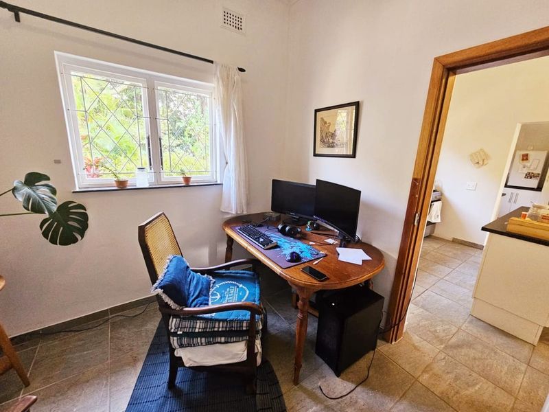 A functional home office space featuring tiled flooring, a window with security bars, and direct access to the adjacent kitchen area.
