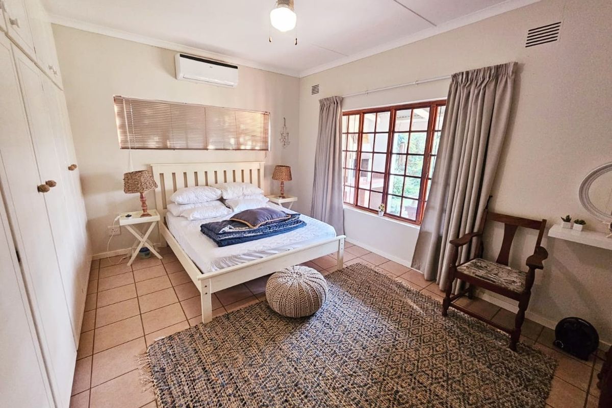 A bedroom featuring tiled flooring, built-in cupboards, a ceiling fan, and a wall-mounted air conditioning unit with timber-framed windows.