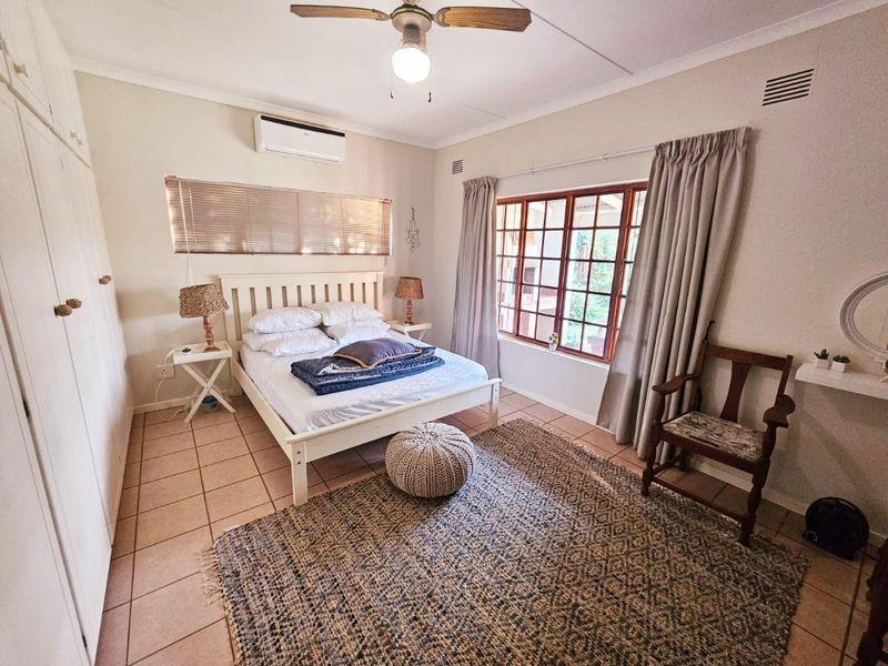 A bedroom featuring tiled flooring, built-in cupboards, a ceiling fan, and a wall-mounted air conditioning unit with timber-framed windows.