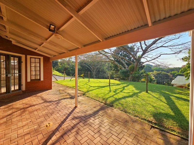 A covered brick-paved patio overlooking a spacious, fenced garden area with mature trees and a scenic outlook.