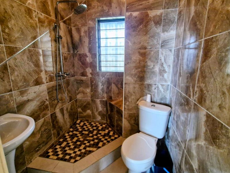 A compact bathroom featuring floor-to-ceiling brown marble-effect tiles, a corner shower enclosure with a mosaic tile floor, a pedestal basin, and a toilet.