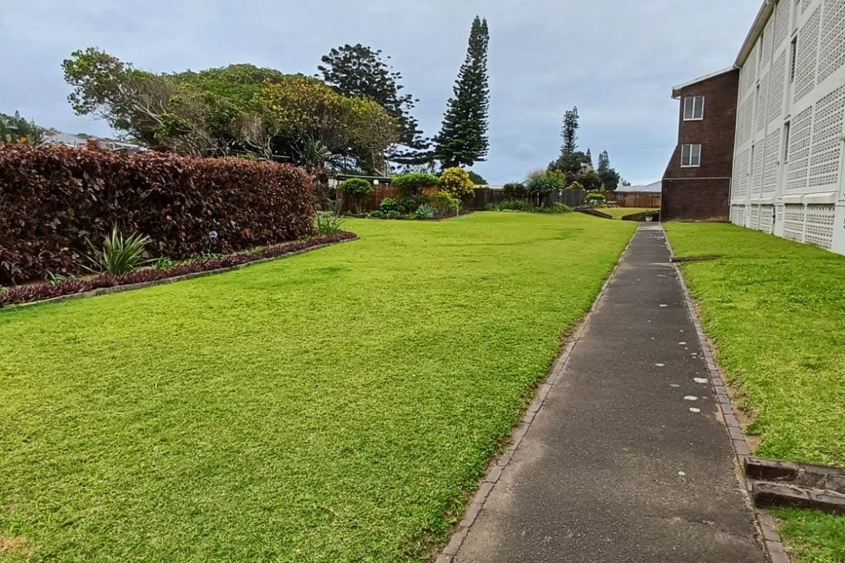 A well-maintained rear garden area featuring a paved walkway alongside the apartment building, bordered by a mature hedge and landscaped lawn.