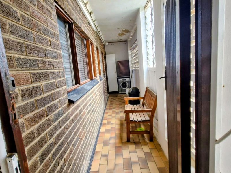 A covered balcony or walkway featuring brick walls, tiled flooring, and louvered windows, showing signs of wear on the ceiling.