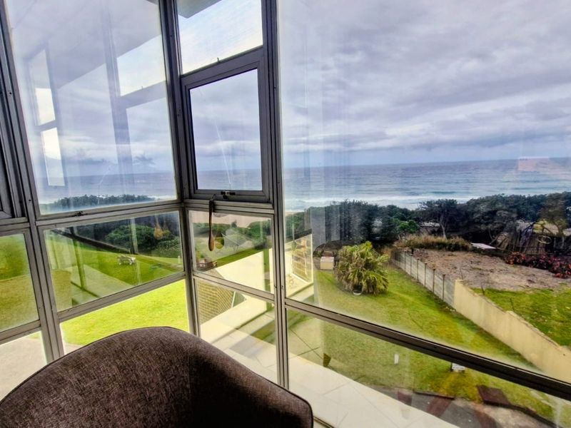 A bright enclosed balcony or sunroom featuring expansive glass windows offering panoramic views of the ocean and surrounding coastal landscape.