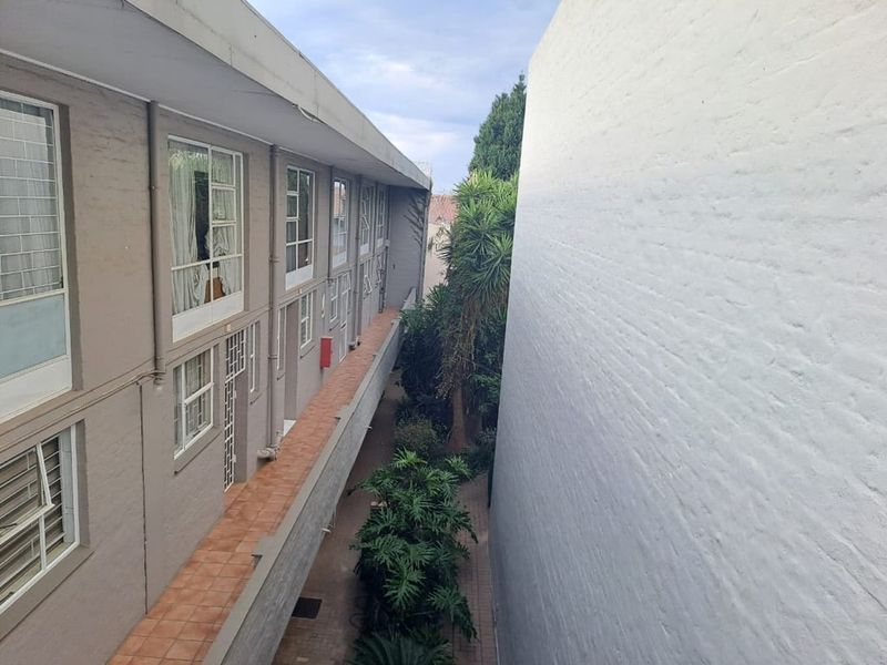 Exterior view of the apartment building showing a tiled walkway, brick facade, and metal window frames overlooking a landscaped courtyard area.