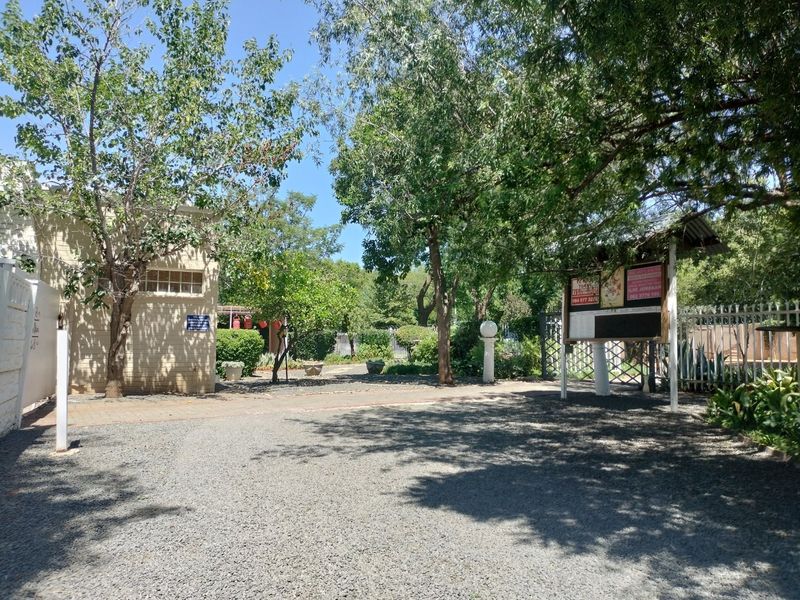 A gravel driveway entrance featuring mature trees, established landscaping, and perimeter security fencing.