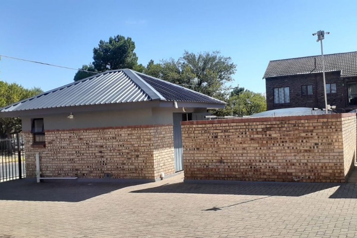 A view of a detached outbuilding featuring a metal roof and facebrick exterior, situated on a paved property with security fencing.